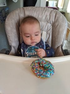Baby stares down at donut with blue frosting and rainbow sprinkles. Some donut is in his fist.