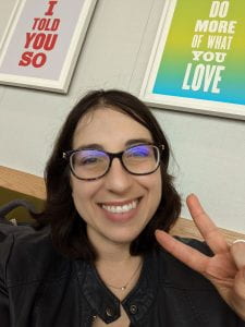 A smiling brunette woman faced the camera and holds up a peace sign. Behind her are two art prints in block letters that say "I TOLD YOU SO" and "DO MORE OF WHAT YOU LOVE."